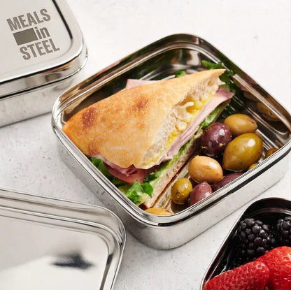 A stainless steel square lunch box filled with a sandwich, olives, and greens, shown on a light stone surface with other matching containers partially visible. The lid has the text 'MEALS in STEEL' printed on it.

