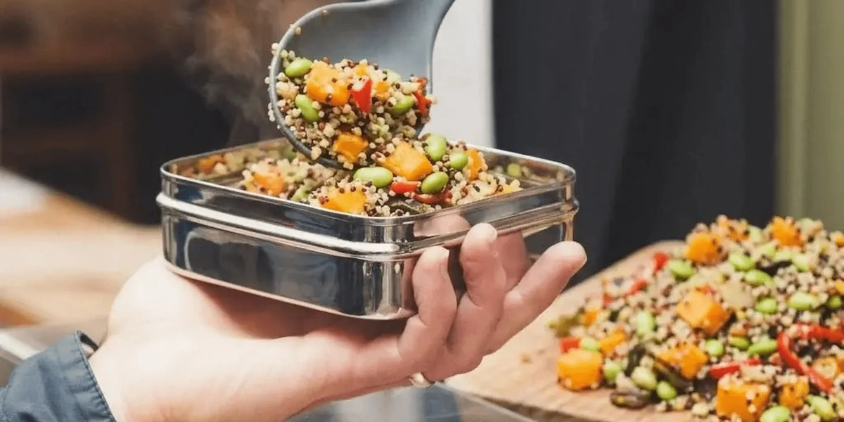 A stainless steel lunch box being held in a hand, with a spoon lifting a colorful grain salad above it. More of the salad is visible on a board in the background.